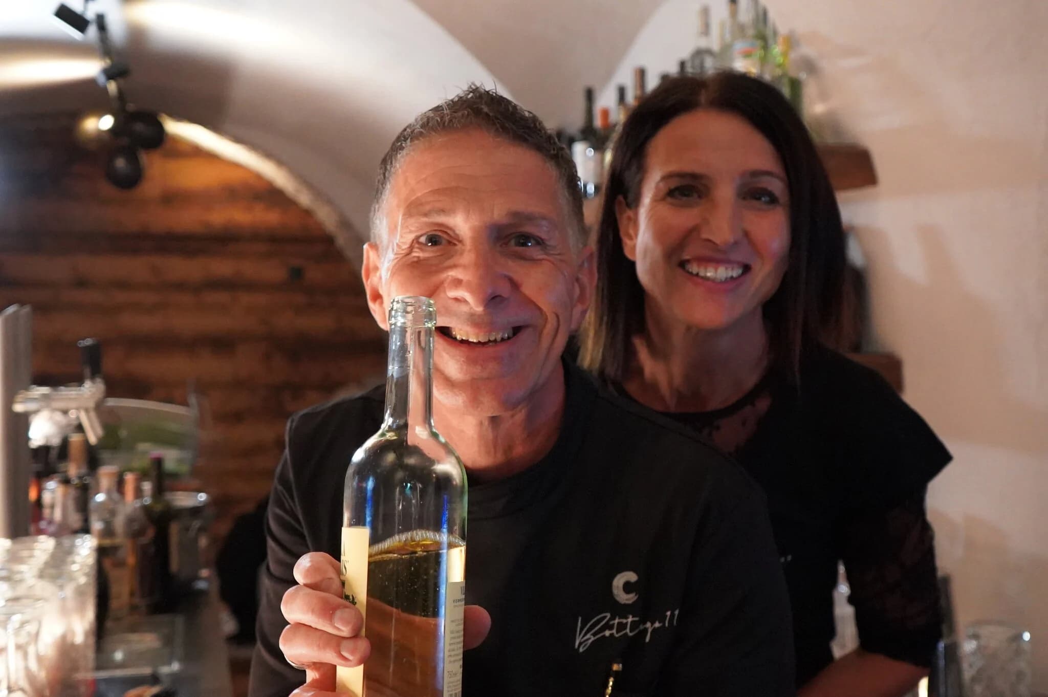Massimiliano und Susanne Arioni lachend hinter der Bar, er hält eine Flasche Vermentino